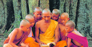 6 buddhist monk children surrounding an adult reading from a book with tree trunks in background
